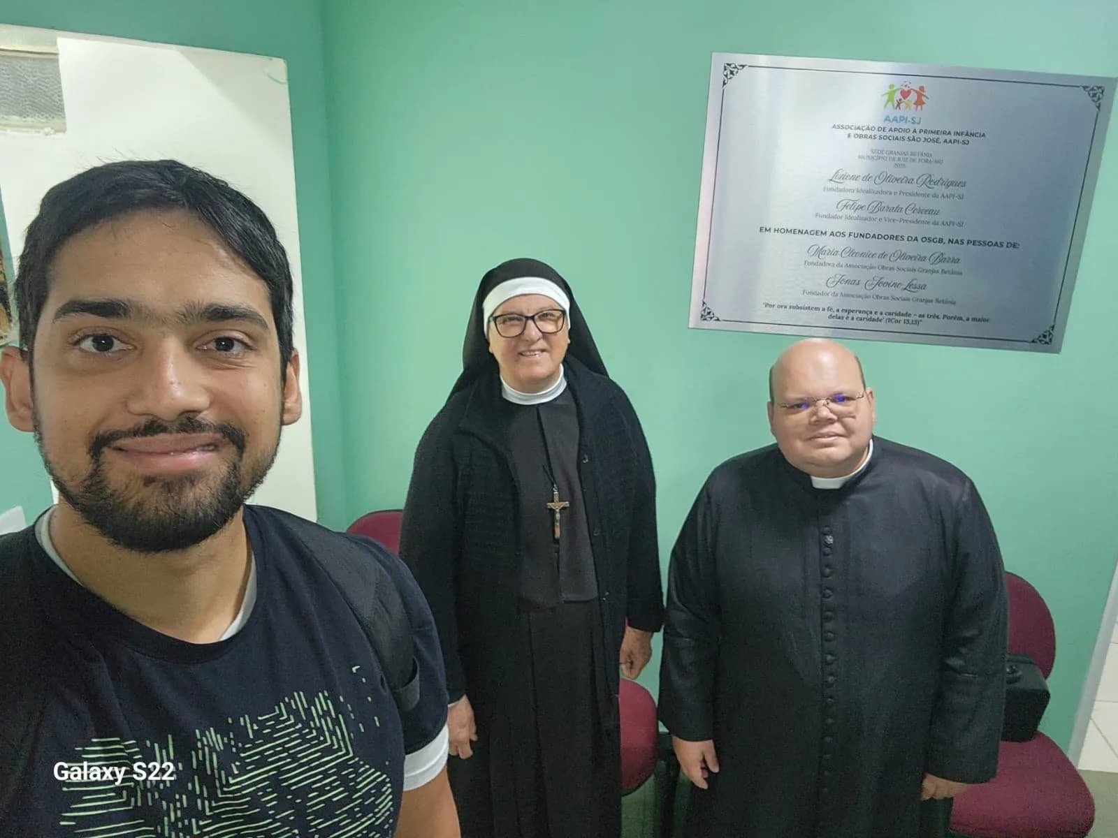 Selfie com o Padre Jean Marcos e a irmã religiosa ao lado da placa inaugural da AAPI-SJ